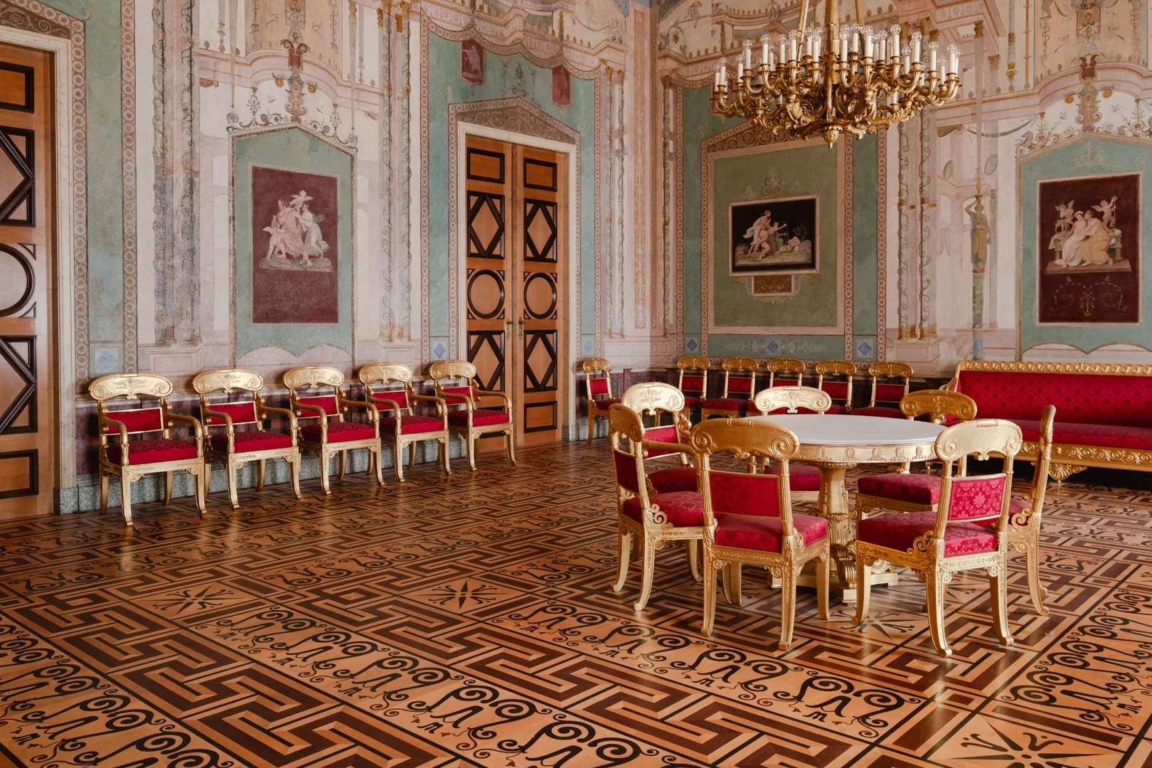 An ornate, historic room with a decorative wooden patterned floor, elegant chairs with red and gold upholstery arranged around a white circular table, and similar chairs in a line against the wall. The walls are decorated with classical paintings and