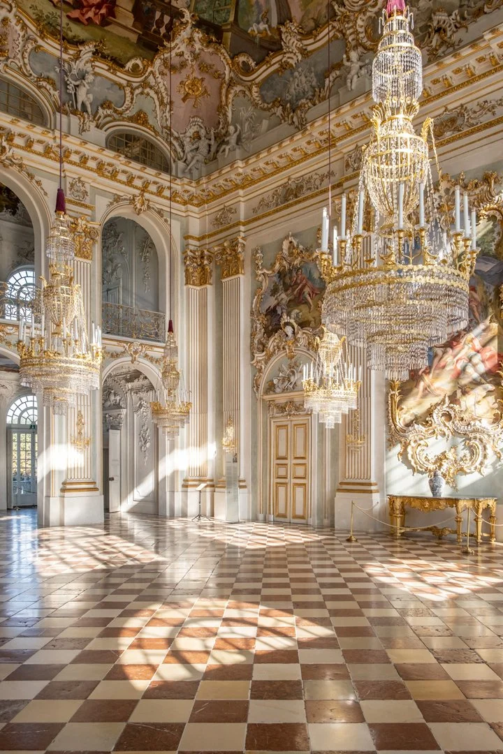 Opulent palace hall featuring ornate gold and white decor, large chandeliers, intricate wall and ceiling murals, and checkered marble floors.