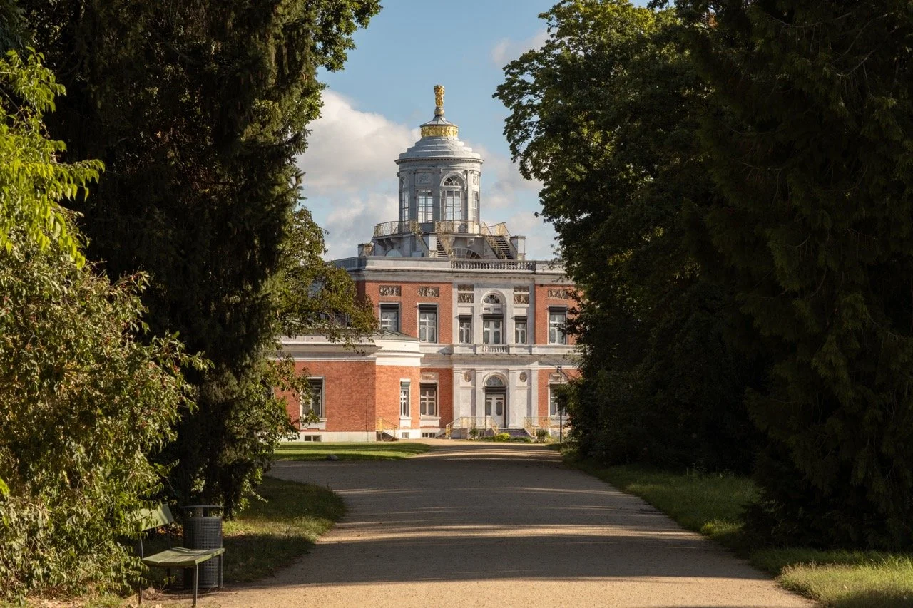 A historic building viewed from a pathway surrounded by trees, with a golden statue on top and a partly cloudy sky in the background.