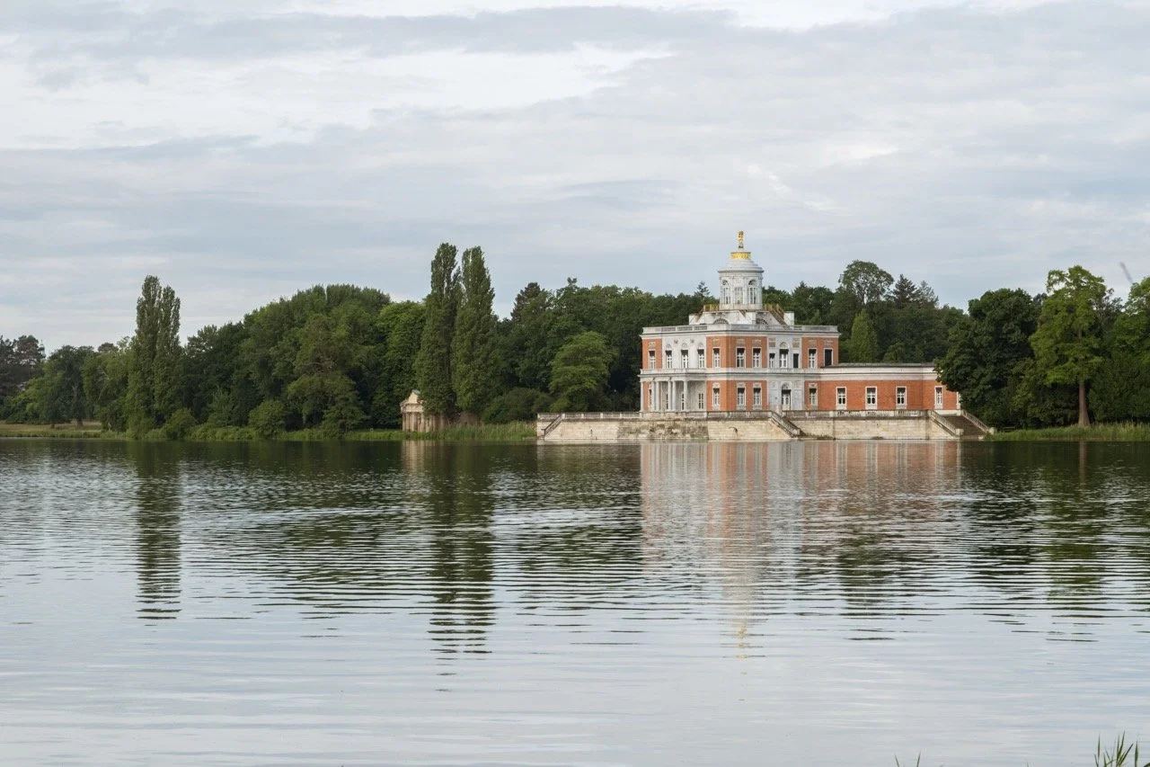 A historic building with a statue on top, situated on the edge of a lake, surrounded by trees, under a cloudy sky.