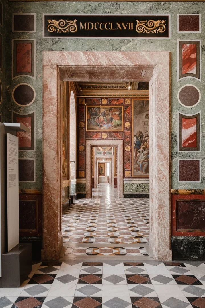A series of doorways creating a corridor with ornate brick and marble walls, decorated with classical paintings and intricate patterns, in a historic building.