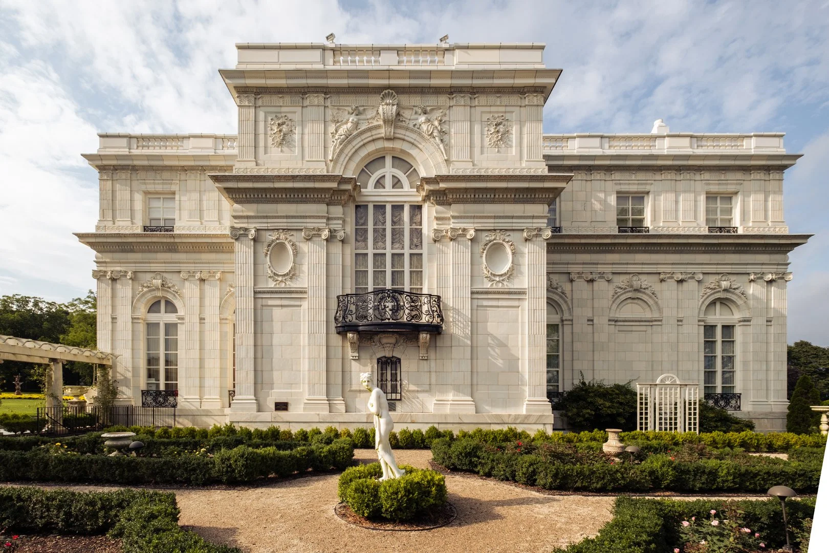 Front view of a grand white mansion with intricate architectural details, tall windows, and a small balcony. A garden with a statue and manicured bushes is in front of the building.