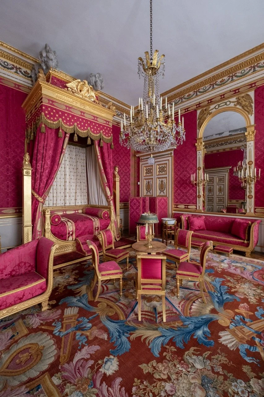 An ornate room with red and gold decor, including pink velvet chairs, a pink velvet sofa, a four-poster bed with pink curtains, a chandelier, large mirror, and intricate patterned carpet.