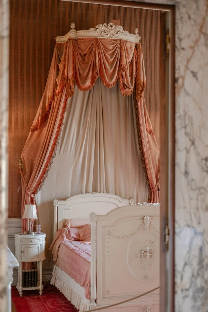 A vintage bedroom with a white wooden bed, pink bedding, and an elaborate canopy with pink and peach drapes, ornate detailing, and a marble wall partially visible.