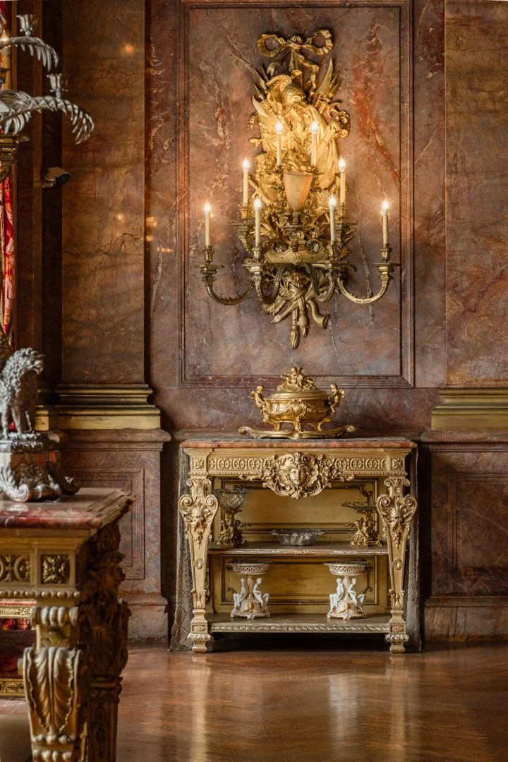 An ornate, gold-colored wall sconce with candles, mounted on a richly decorated marble wall with a decorative console table beneath it, in an elegant, classical interior setting.