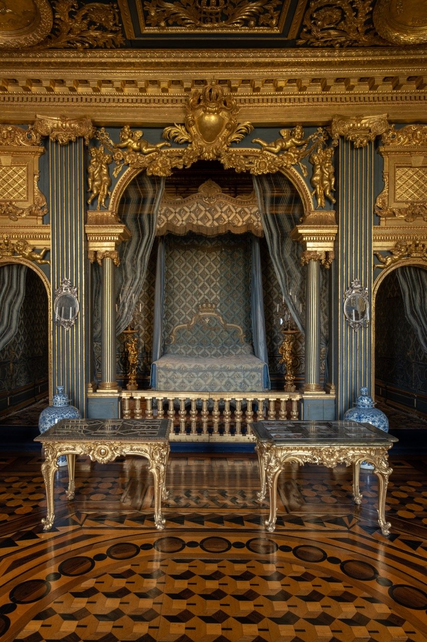 Ornate royal bedroom with a blue and gold color scheme, featuring a canopy bed with a patterned bedspread, gold detailing, and draped curtains, surrounded by decorative tables and classical wall decorations.