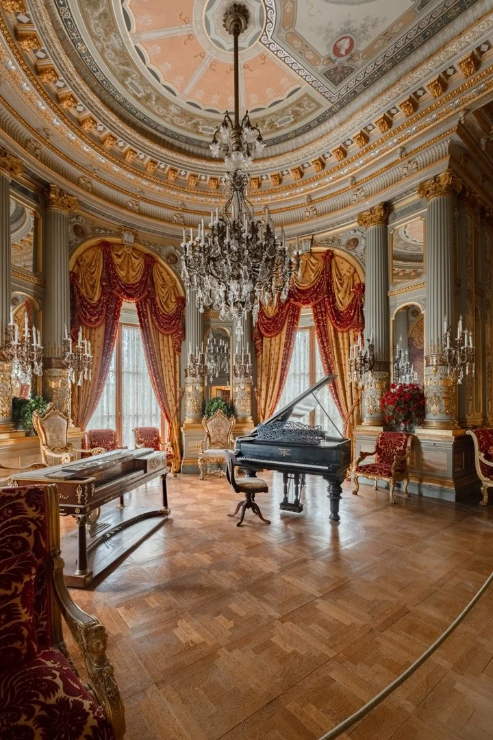 Elegant and luxurious room with a grand chandelier, a black grand piano, ornate gold-trimmed furniture, large windows with red and gold curtains, and intricate ceiling designs.