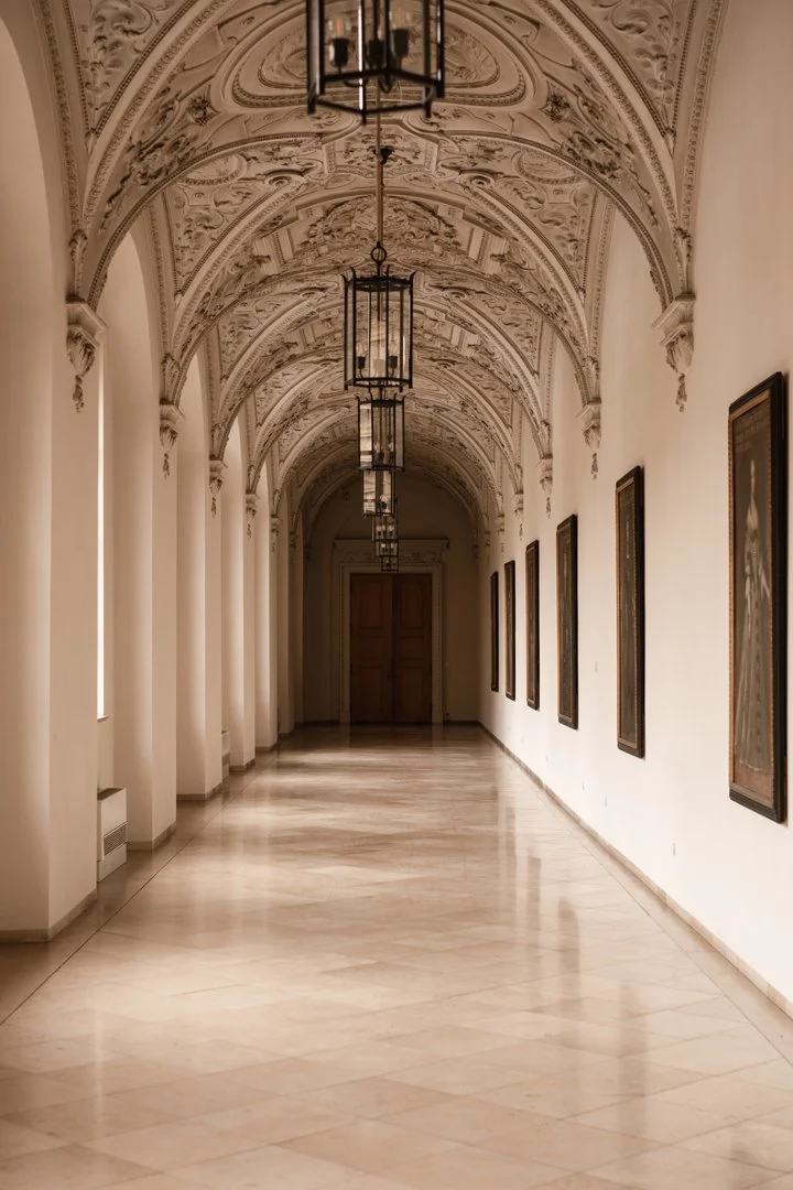 Elegant hallway with intricate ceiling design, hanging lantern-style lights, framed artwork on the right wall, and tall windows on the left, leading to a wooden door at the end.