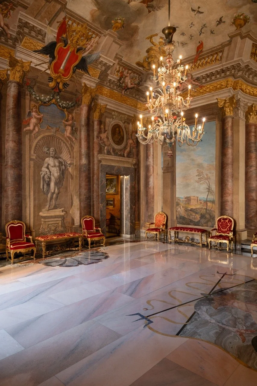 An opulent room with elaborate mural paintings on the walls and ceiling, featuring classical sculptures, cherubs, and a landscape scene. The room is decorated with red velvet armchairs, gold accents, and a large chandelier hanging from the ceiling.