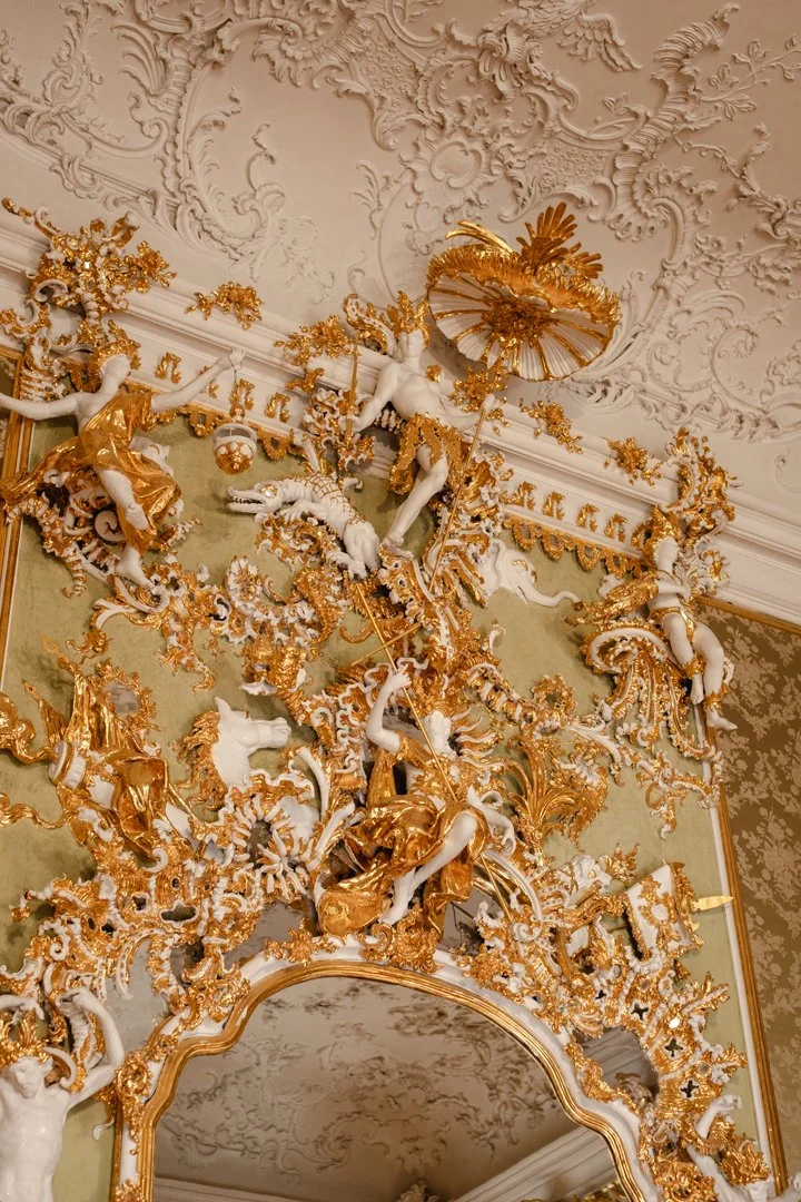 Ornate ceiling and wall decor with gold and white sculptures of cherubs and mythological figures, elaborate gold floral accents, and a decorative mirror frame