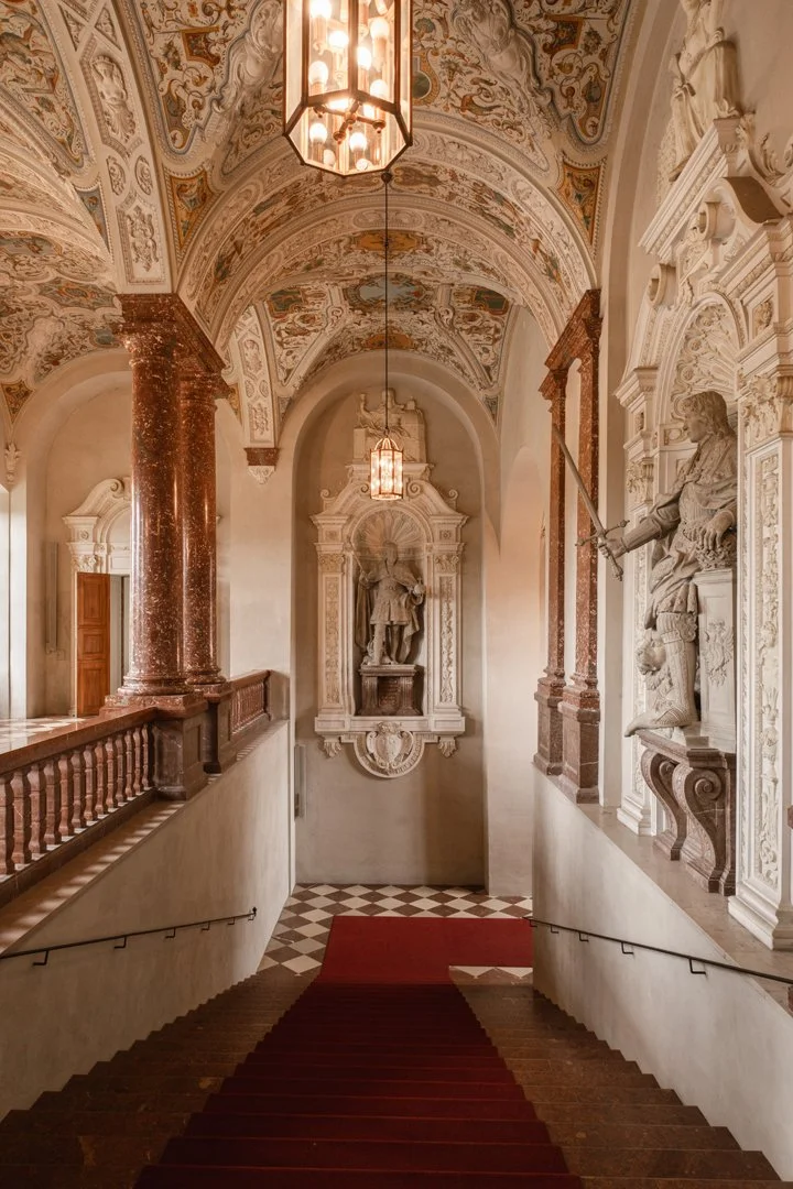 Elegant staircase with a red carpet leading down, ornate ceilings with detailed paintings, and statues of historical figures in decorative alcoves along the walls.