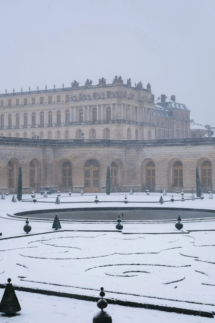 A historic palace or building covered in snow, with a snow-covered courtyard and neatly trimmed trees, under an overcast sky.