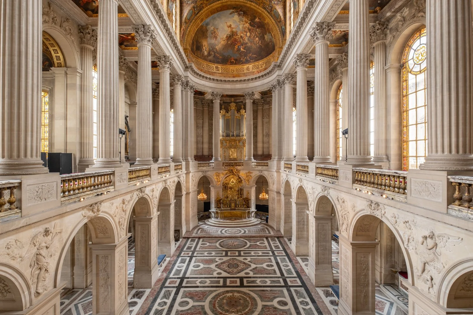 Interior view of a grand, ornate hall with high ceilings, large windows, marble columns, detailed carvings, and an intricate tiled floor.