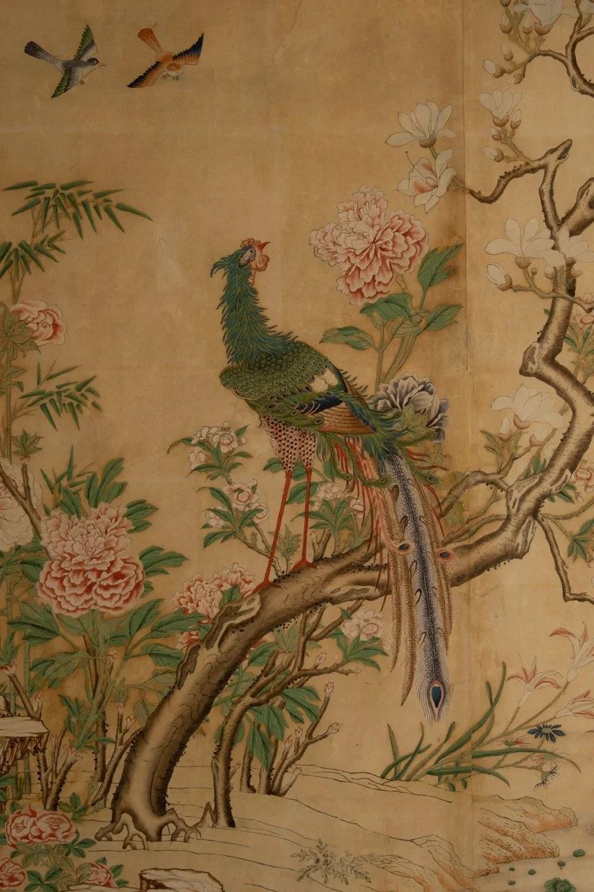 A traditional Asian-style painting featuring a peacock perched on a tree branch surrounded by blooming flowers and two small birds flying above.