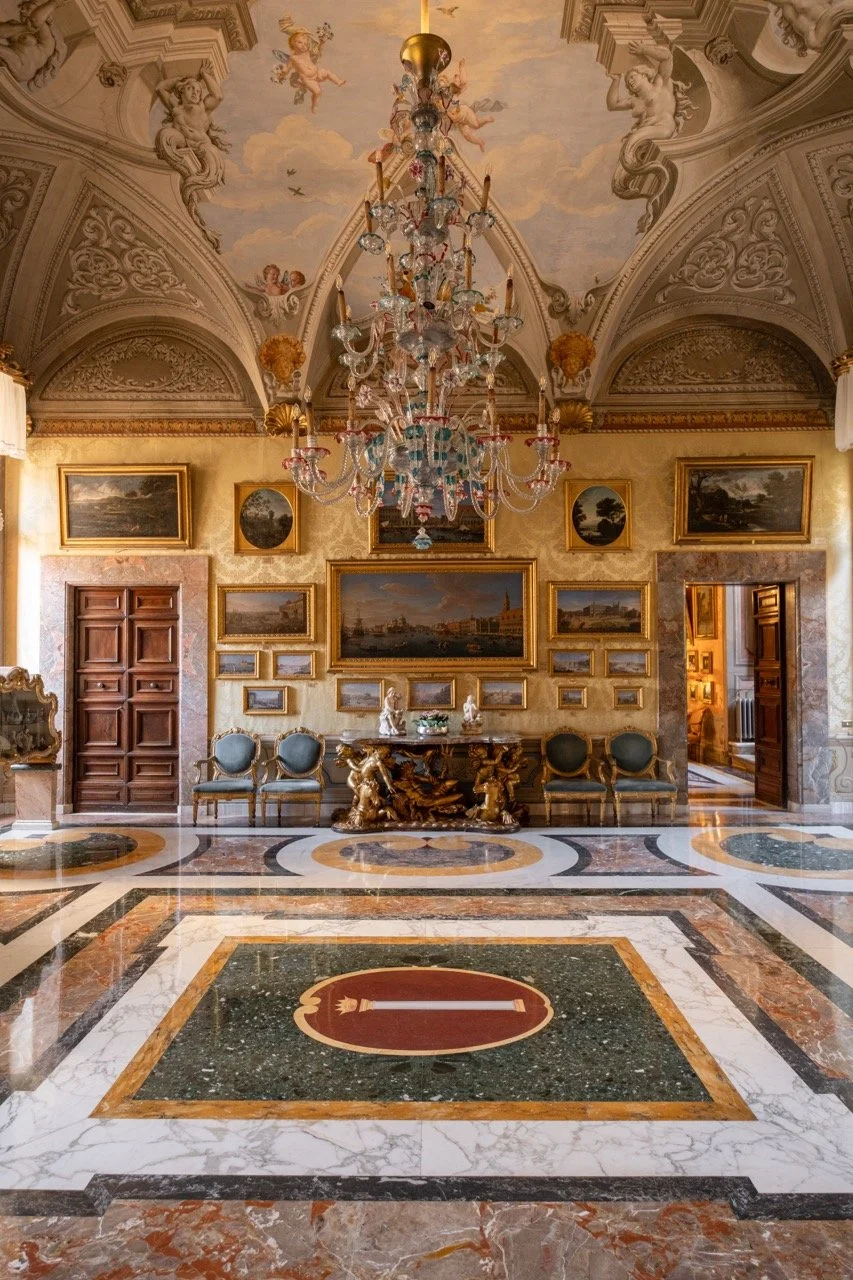 Elegant room with ornate ceilings featuring cherub paintings, a large crystal chandelier, gold-framed paintings and mirrors, a marble floor with geometric patterns, a central red and gold emblem, and a wooden door on each side of the wall.