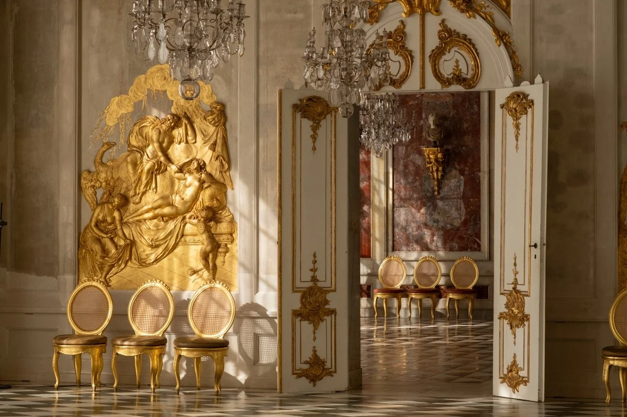 Elegant room with gold and white decor, ornate wall carvings, a large gold relief sculpture on the wall, and matching chairs with round backs arranged in front and inside a doorway. Chandeliers hang from the ceiling, and sunlight casts shadows on the