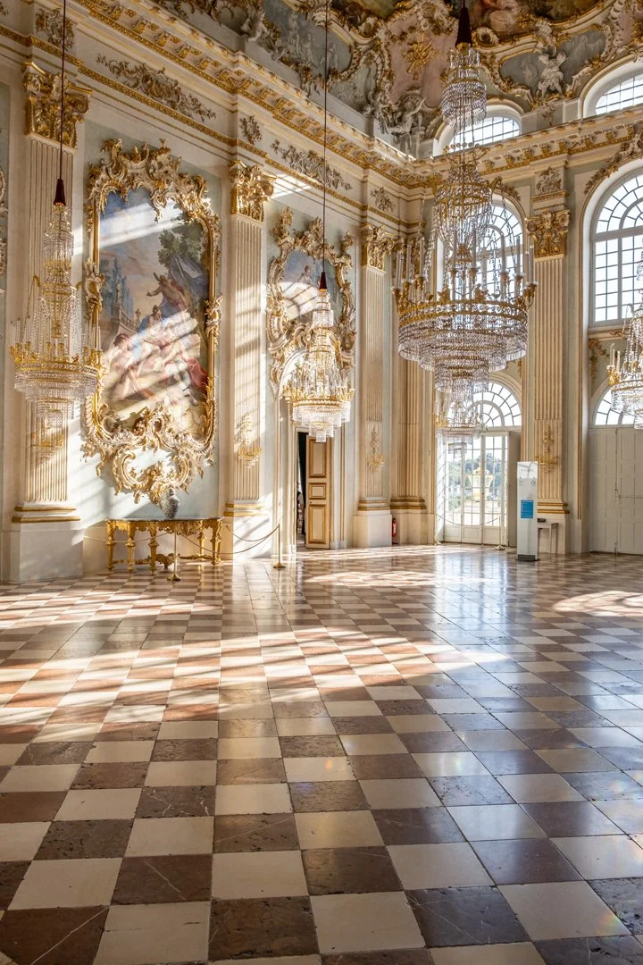 A luxurious, ornate room with high ceilings, large windows, crystal chandeliers, and baroque decor. The floor features a checkered tile pattern, and sunlight streams through the windows, casting shadows on the floor.