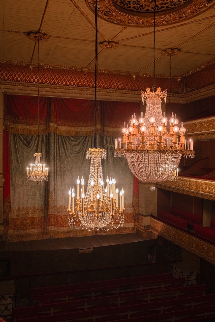 Multiple ornate chandeliers hanging from a decorated ceiling in a theater or opera house, with red velvet curtains and gold detailing.