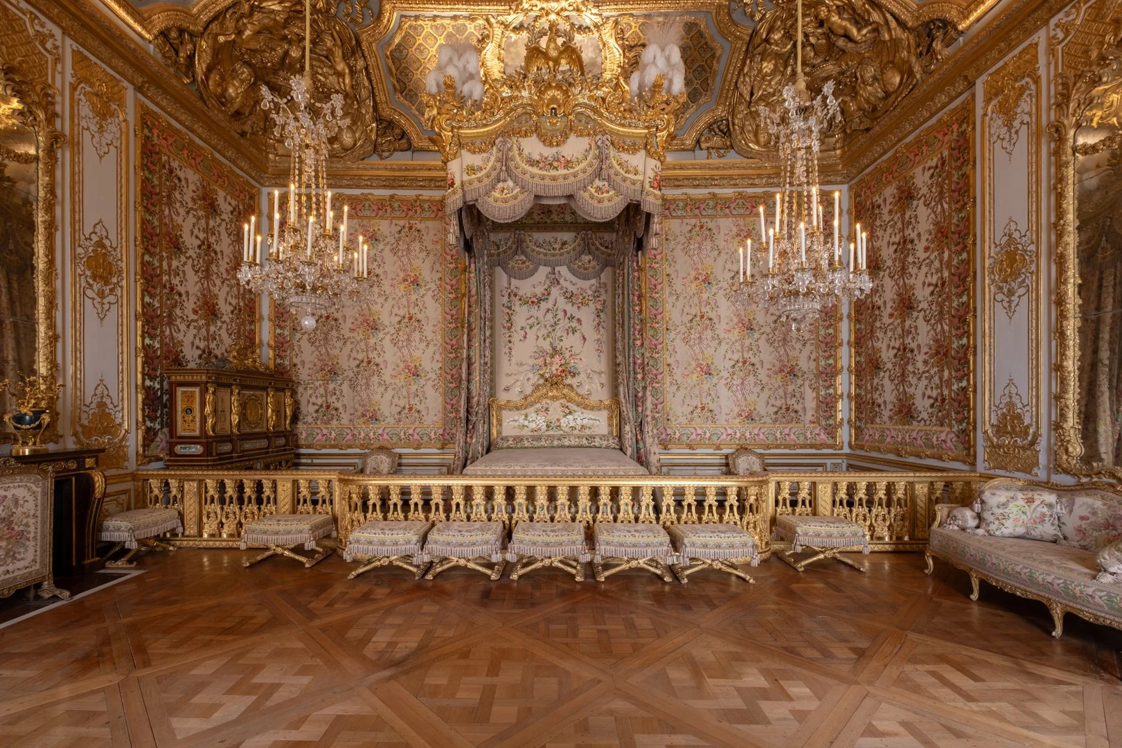 A luxurious, ornate bedroom with gold accents, floral wallpaper, and chandeliers. The room features a canopy bed, several plush stools, and elegant furniture.