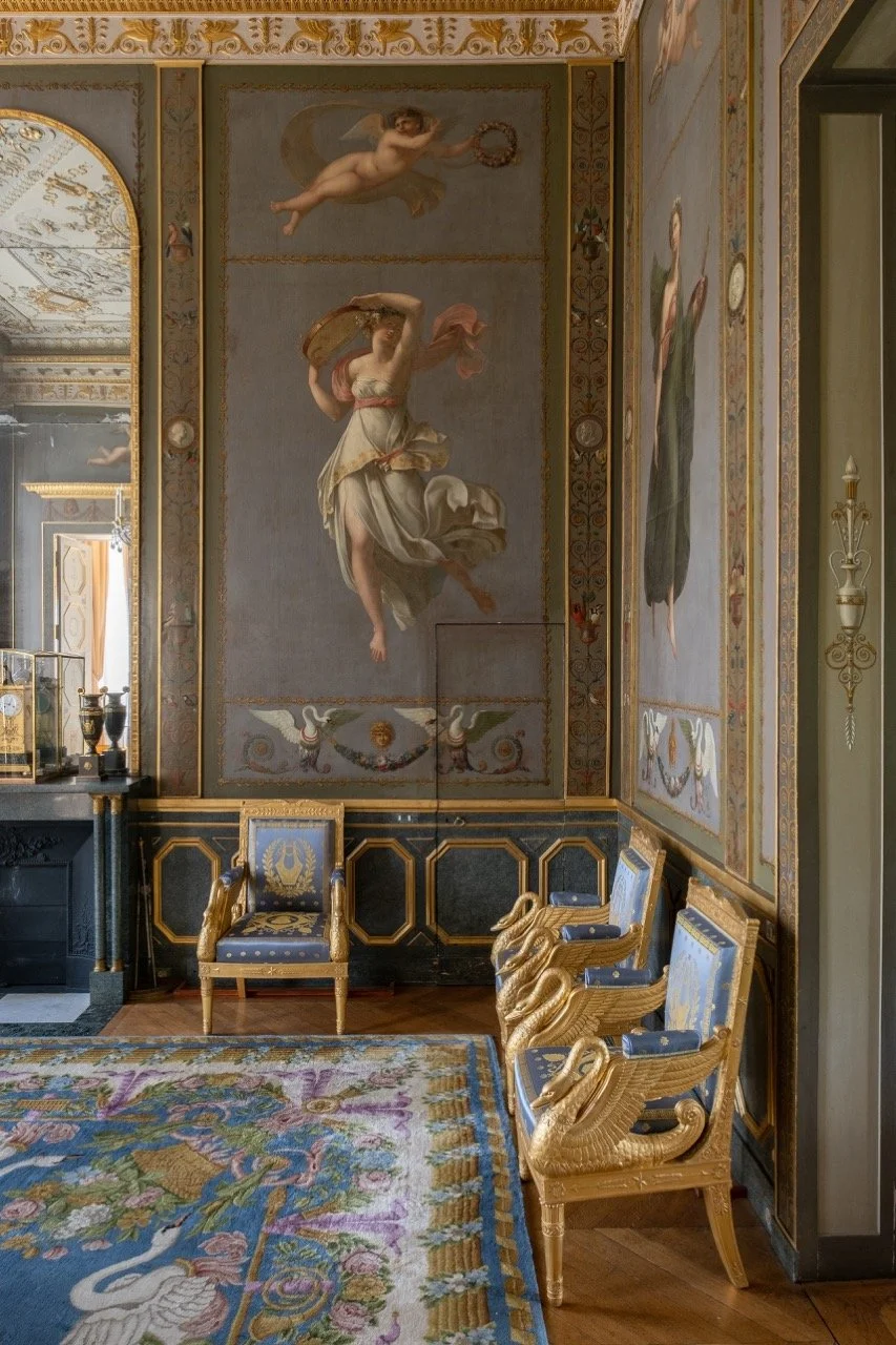 Luxurious room with ornate furnishings including gold-accented chairs with swan motifs, a colorful floral rug, and large classical-style wall murals depicting mythological figures and decorative elements.