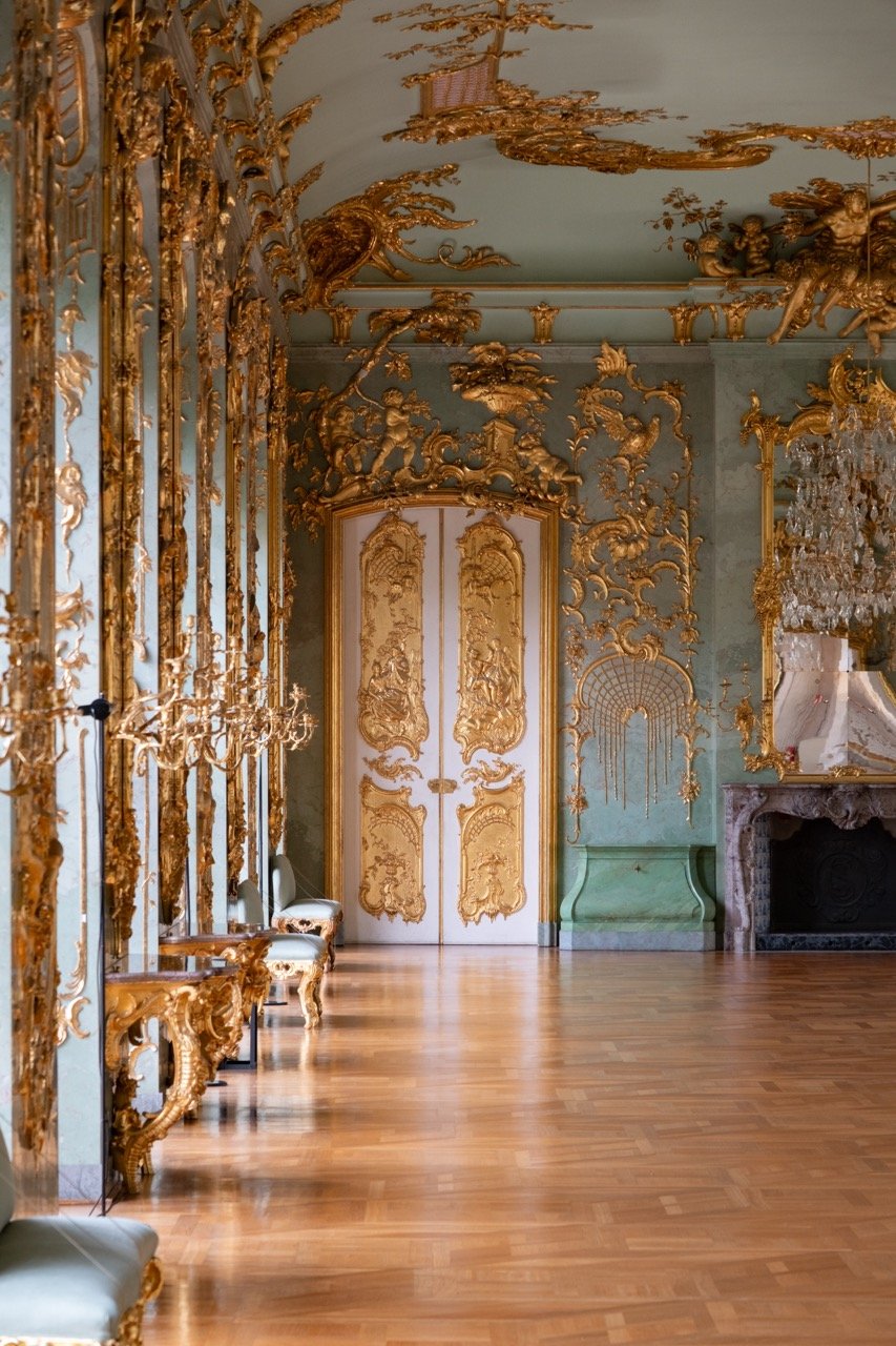 An opulent room with gold ornate decorations on the walls and ceiling, light-colored wooden floor, and elegant furniture.