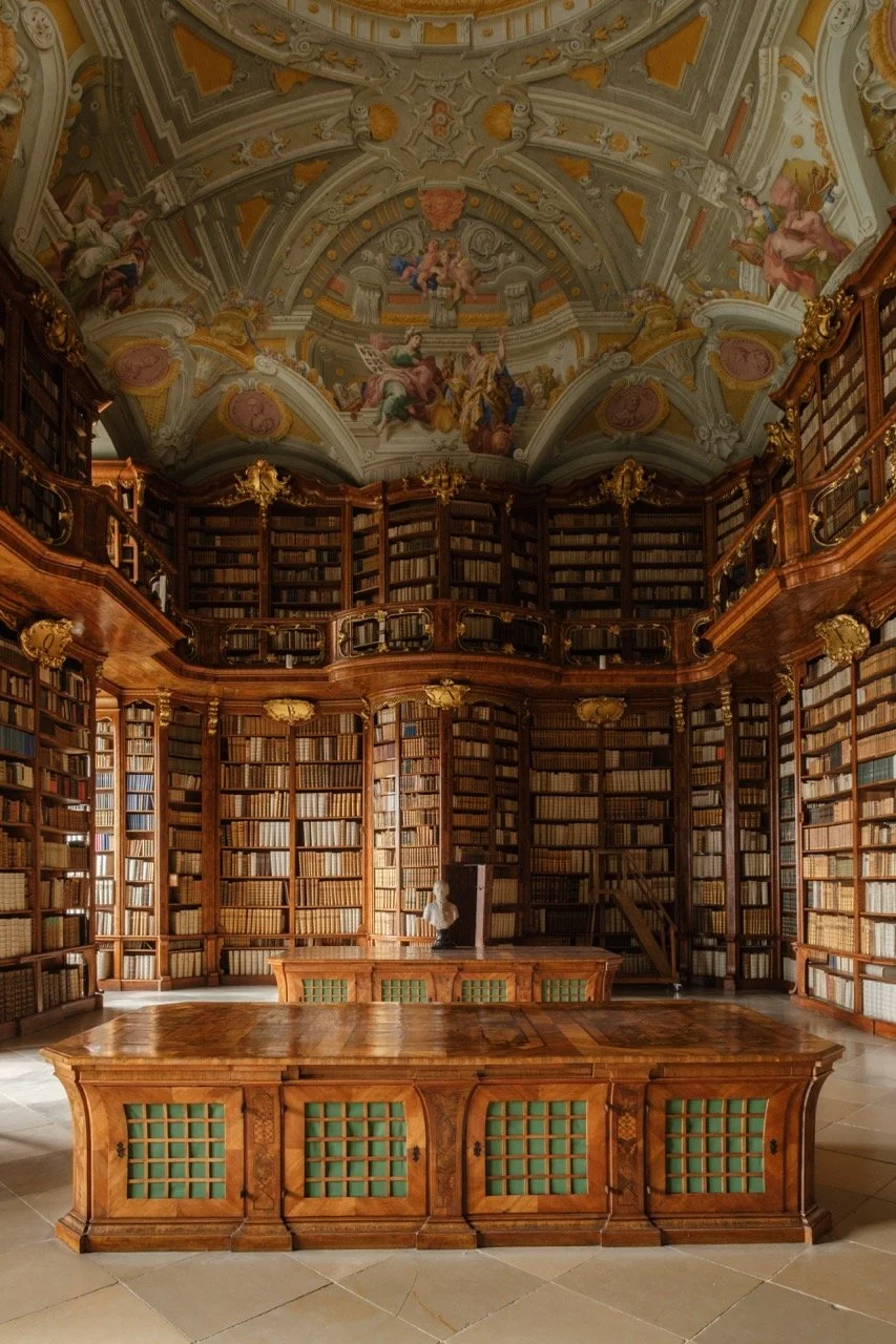 An opulent library with ornate wooden bookshelves filled with books, a high ceiling with intricate frescoes and gold accents, and a large wooden table in the center.