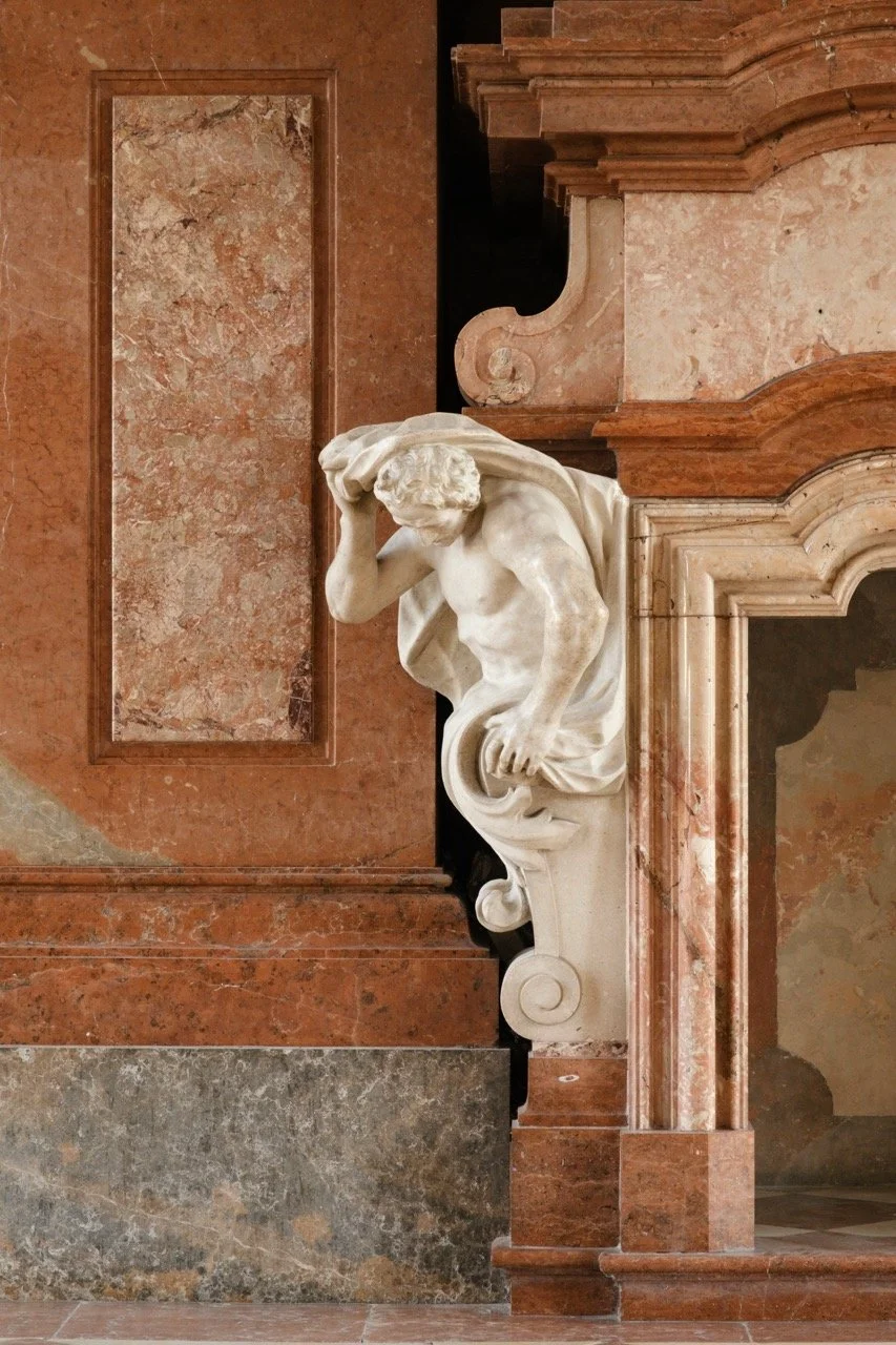 A classical marble sculpture of a muscular male figure with a cloth draped over his shoulder, set against a marble and stone wall with ornate architectural details.