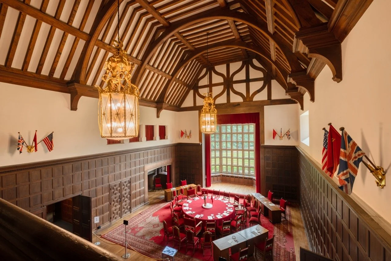 An elegant meeting room with high vaulted wooden ceiling, red curtains, a large round table with red chairs, flags on the walls, and large windows letting in natural light.