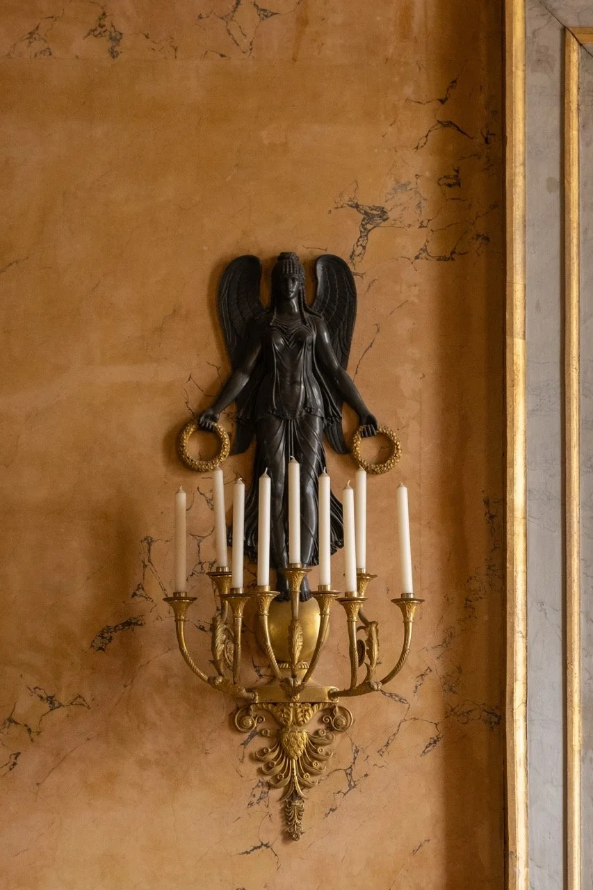 Statue of a winged female figure holding a wreath, mounted above a gold-colored candelabrum with seven white candles, on a marble wall.