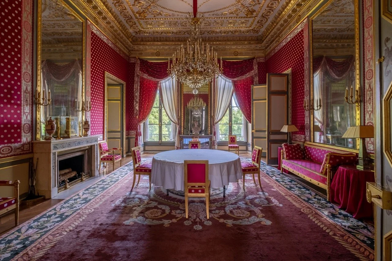 A luxurious, ornate room with red and gold decor, a chandelier, large windows with draped curtains, a fireplace, a round table with a white tablecloth, and elegant chairs and sofas.