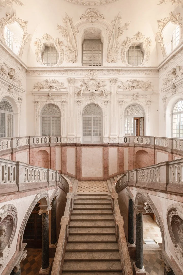 Interior view of an ornate, classical-style grand staircase in a historic building with detailed white carvings, large arched windows, and a domed ceiling.
