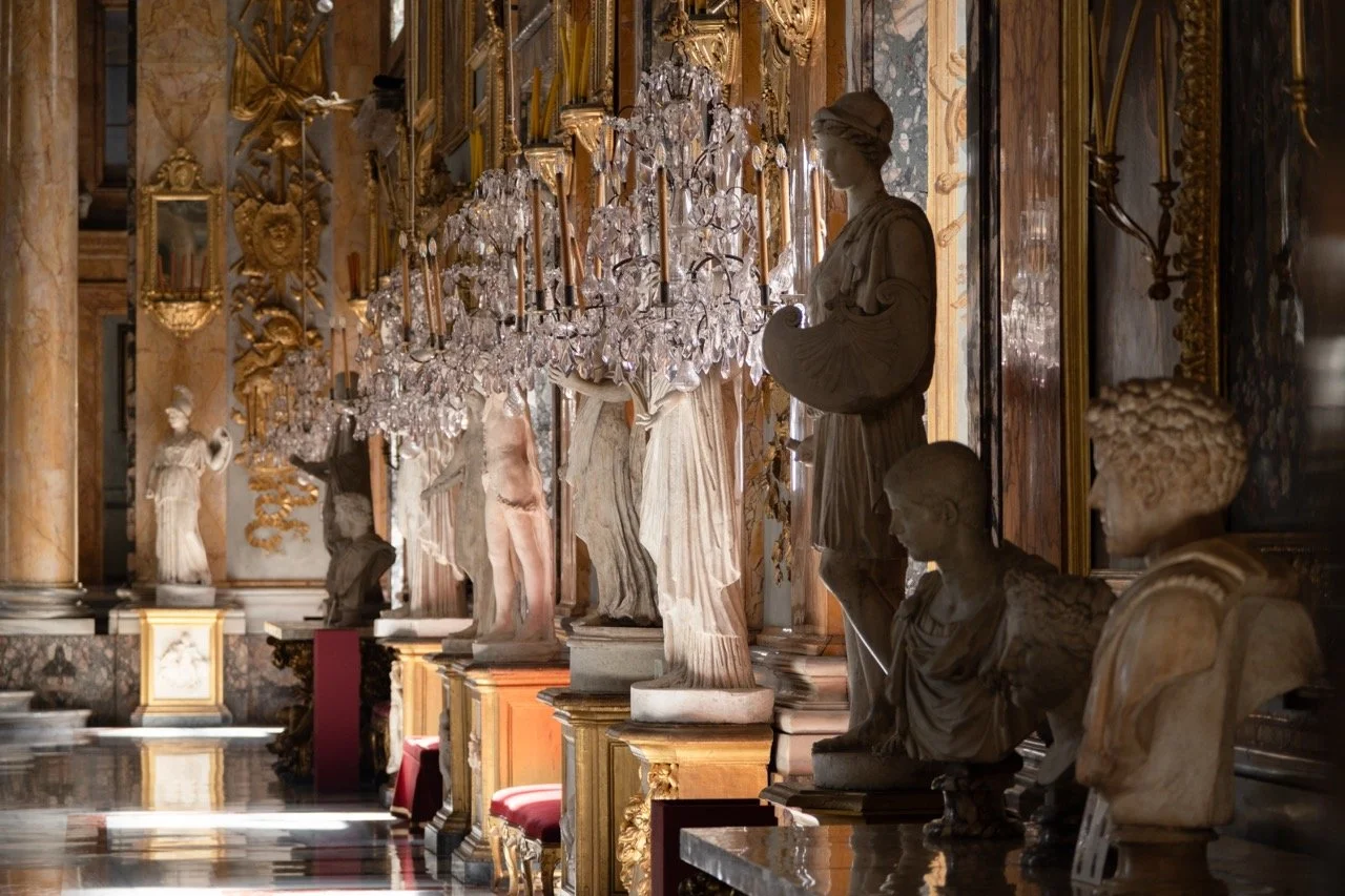 A grand hall with marble statues lining the wall, ornate gold accents, and a large crystal chandelier hanging overhead.