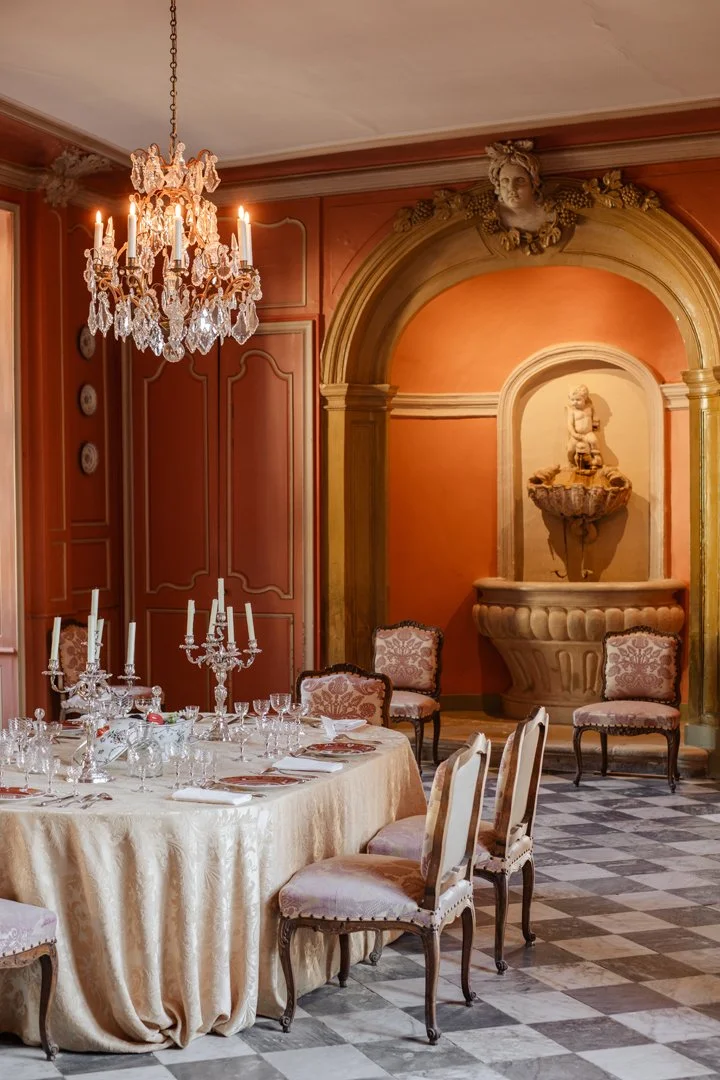 Elegant dining room with a large round table covered in a cream tablecloth, set with glassware, candles, and silverware. There is a crystal chandelier hanging from the ceiling, pink walls with decorative trim, and classical chairs with floral upholst