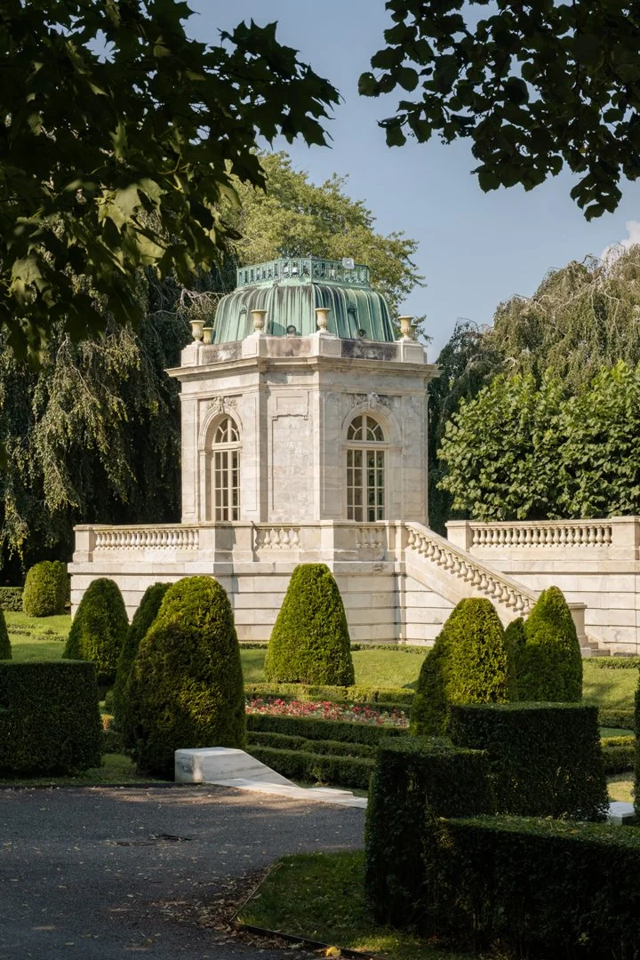 A historic stone building with a green domed roof and large arched windows, situated on a landscaped garden with neatly trimmed bushes and a staircase leading up to it.