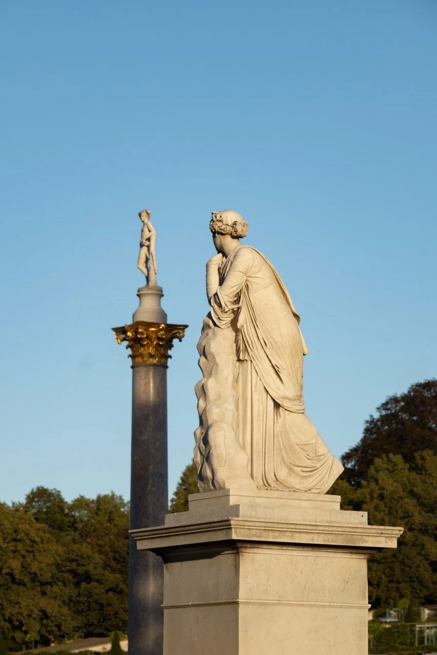 Two classical marble statues, one in the foreground and one in the background on a tall column, set outdoors against a blue sky with trees in the distance.