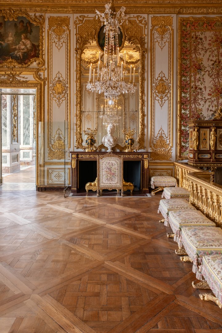 Elegant, gold-accented room with a grand chandelier, ornate mirror, marble bust, and floral upholstery furniture, featuring polished wooden floors.
