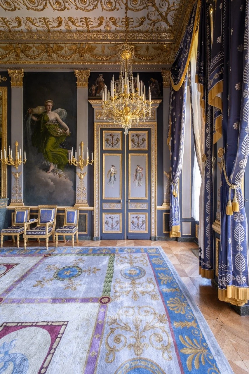 A luxurious, ornate room with blue and gold decor, featuring a gold chandelier, a large painting of a woman in a green dress, and matching blue and gold curtains. There are four blue and gold chairs, a decorative rug, and intricate gold details on th