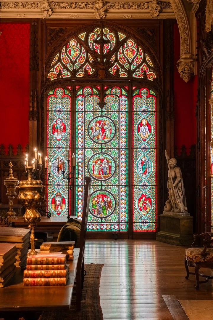Stained glass window with heraldic and religious symbols in a historic, ornate room with antique furniture and statues.