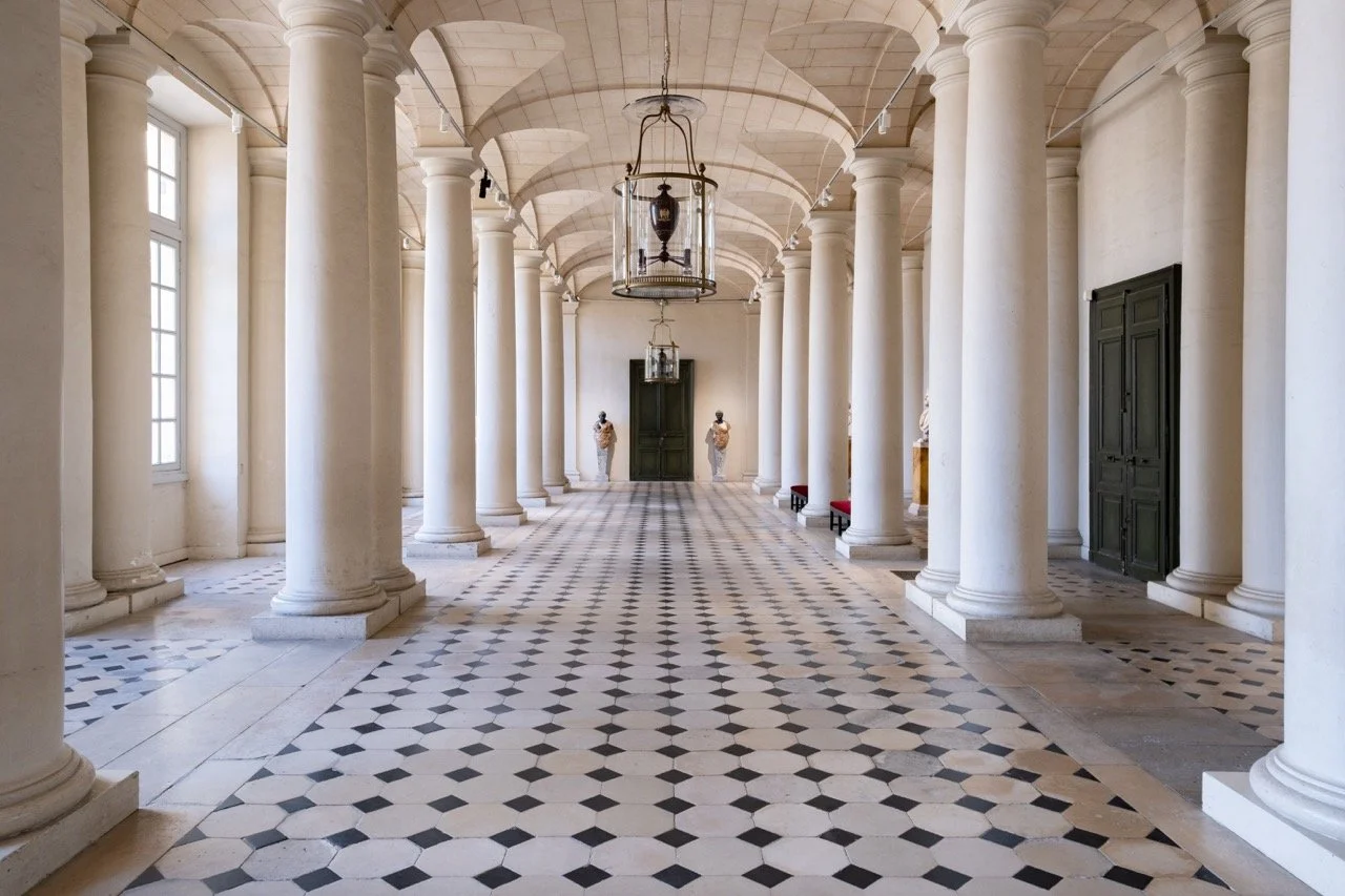 Interior of a grand hallway with tall white columns, checkered floor tiles, large windows on the left, and black doors on the right. There are two statues and hanging lantern lights.