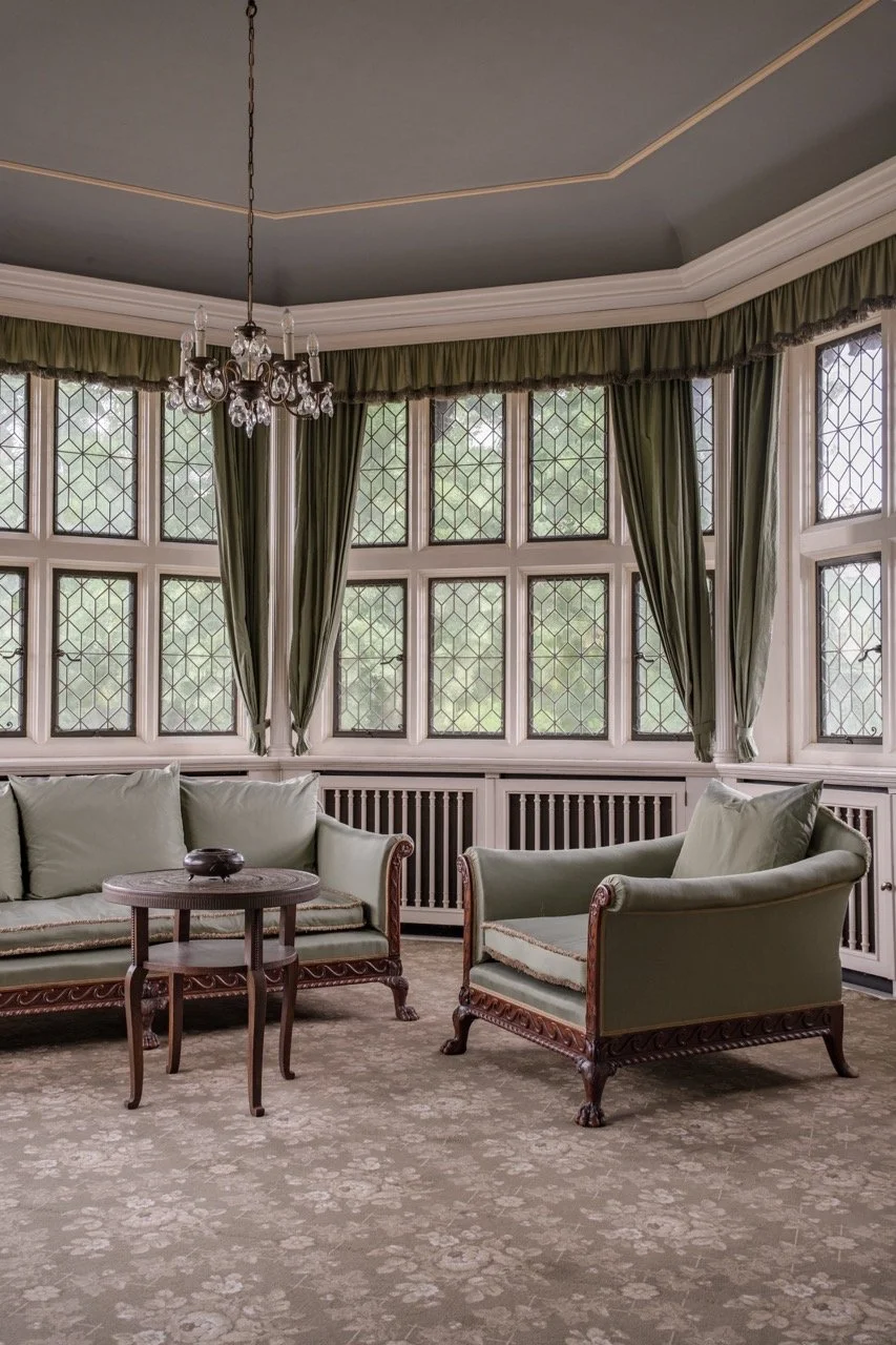 A vintage sitting room with large leaded glass windows, green curtains, a chandelier, a green upholstered sofa, an armchair, a small wooden table, and patterned carpet.