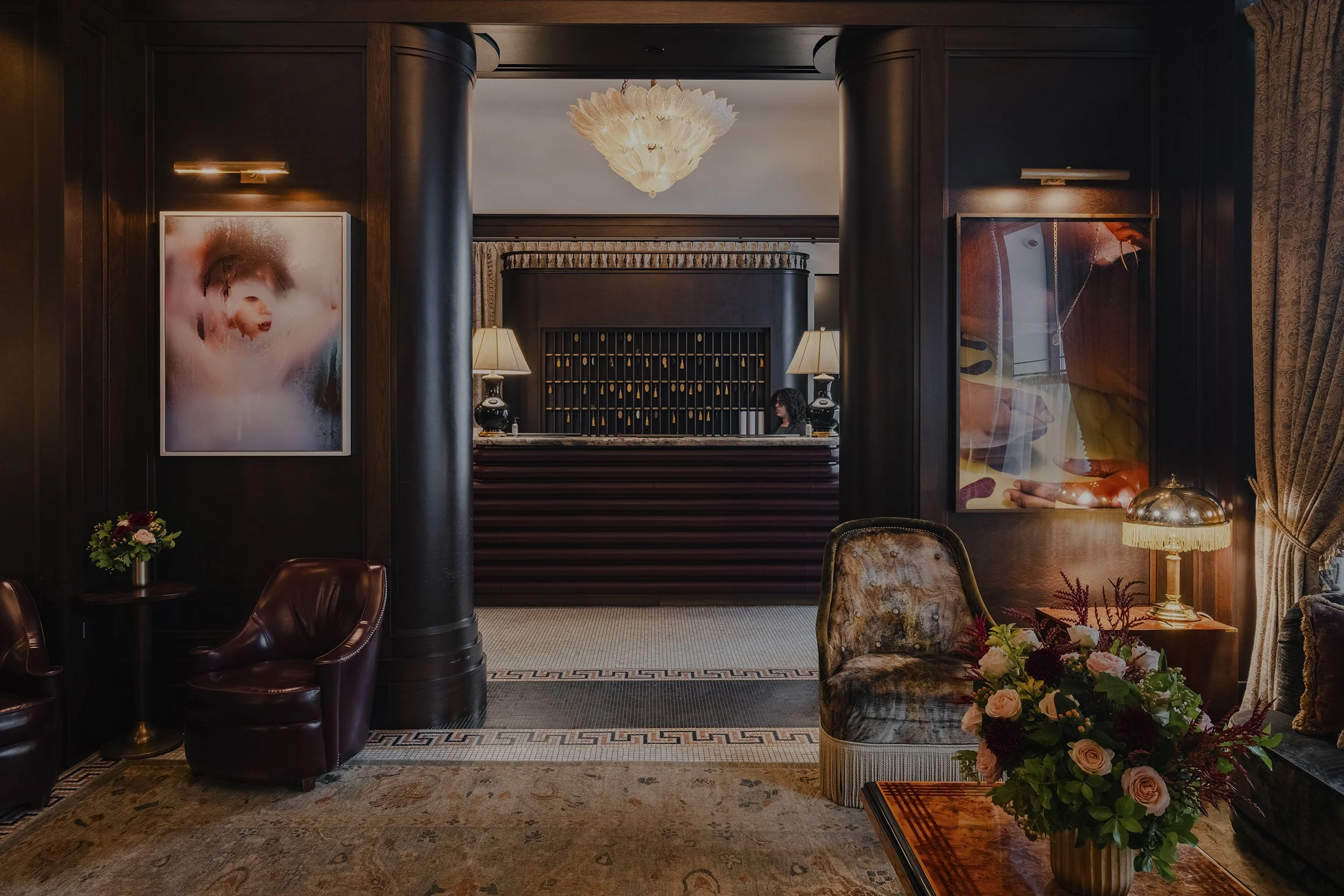 Elegant hotel lobby with dark wood paneling, plush armchairs, floral arrangements, and a curved reception desk with a woman behind it. Two abstract art pieces hang on the walls, and a decorative chandelier hangs from the ceiling.
