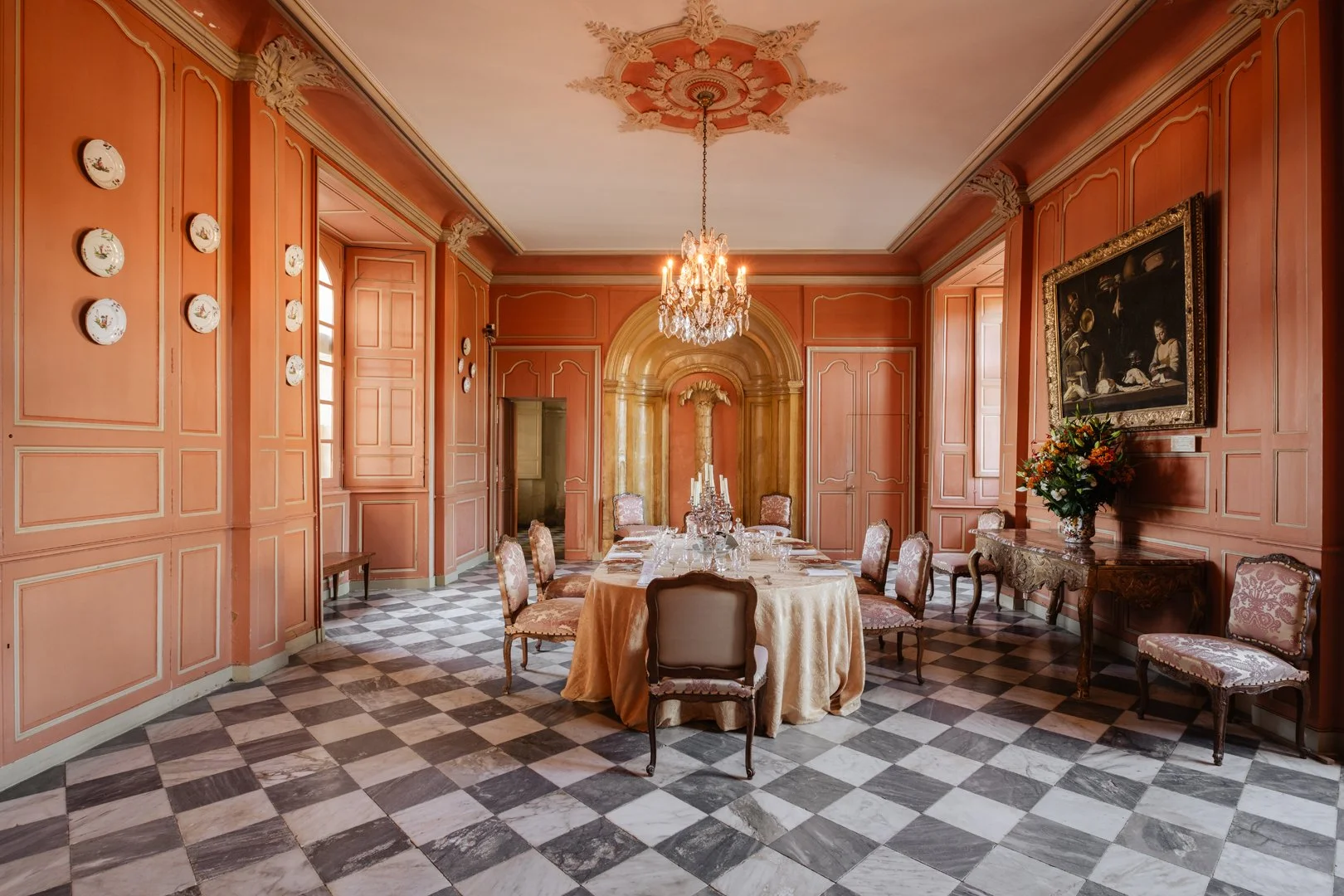 Elegant dining room with pink paneled walls, checkered marble floor, a chandelier hanging from ceiling, a round table with a beige tablecloth, surrounded by ornate chairs, a floral arrangement on a console table, framed artwork on wall, and tall wind