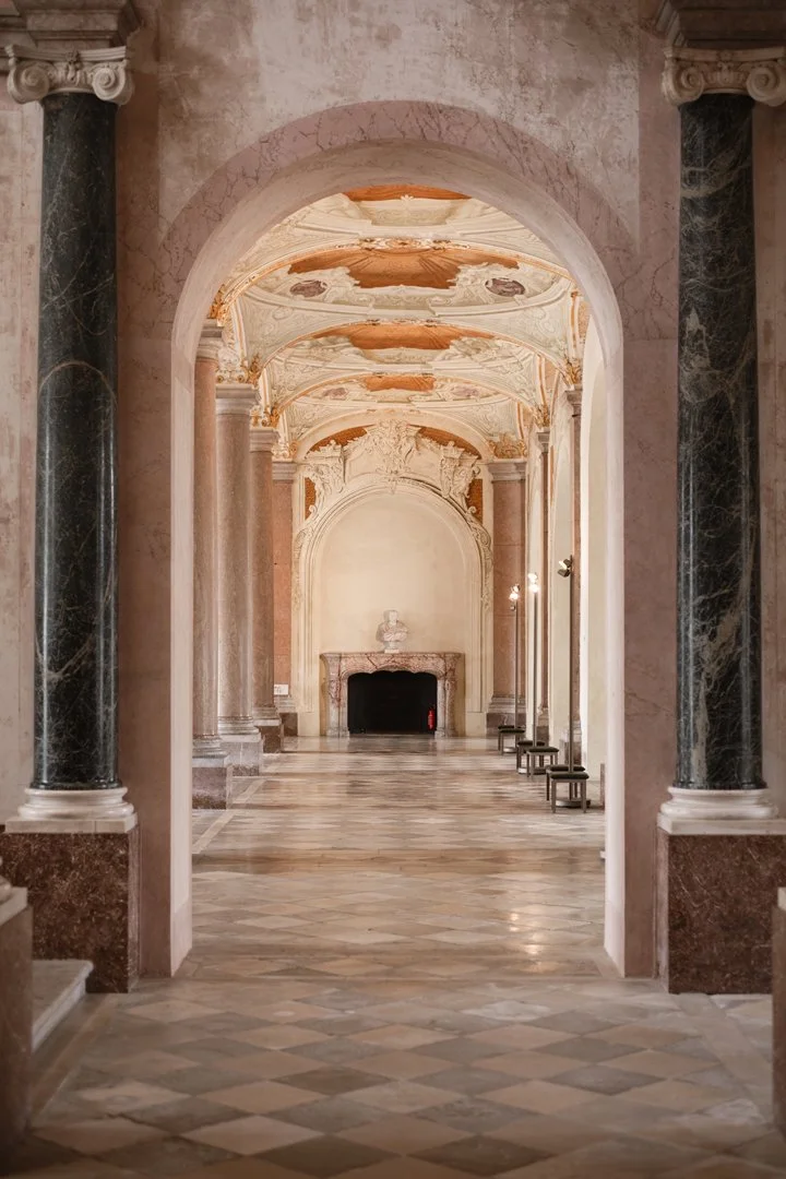 Elegant, ornate hallway with marble columns and intricate ceiling designs, leading to a fireplace with sculpture on top.