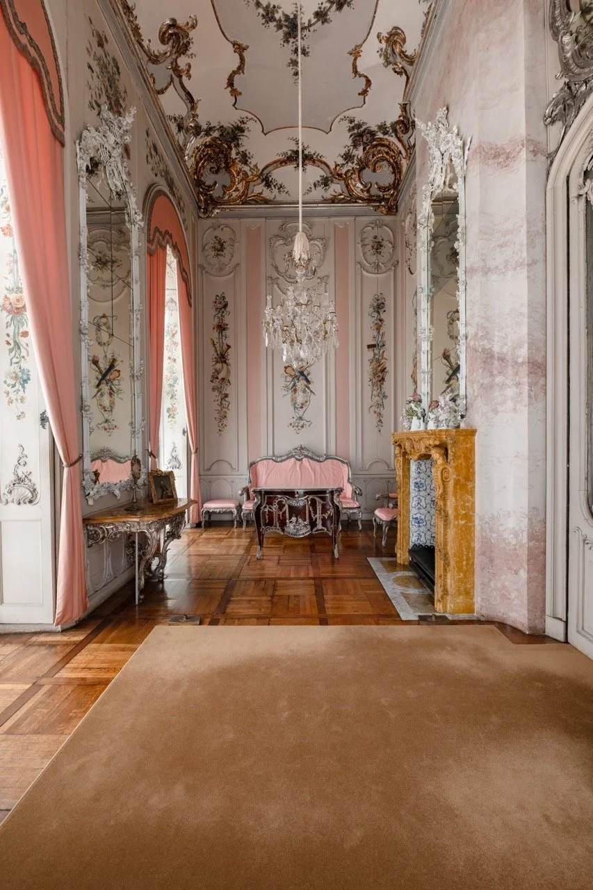 An ornate, historic room with pink and white walls decorated with floral and gilded detailing. There are tall, pink drapes beside large mirrors on the walls. The ceiling features intricate gold and floral patterns, and a crystal chandelier hangs in t