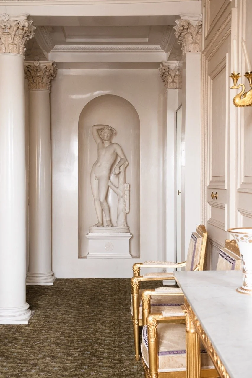 A marble sculpture of a woman standing in a niche, surrounded by classical architectural elements such as columns and ornate moldings, in a luxurious interior setting with a marble-topped table and gold-accented chairs.