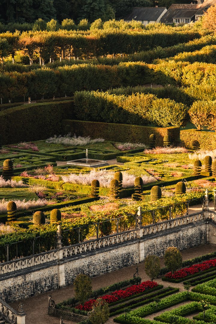 A picturesque garden with well-manicured hedges, flower beds, and a fountain, bordered by a stone wall with decorative railings, set against a backdrop of lush trees and a house on a hill.