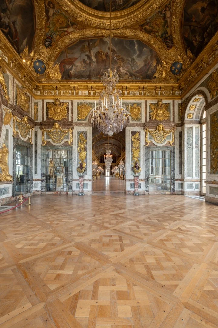 A grand, luxurious palace hallway with a polished wooden floor, ornate gold and marble decorations, large windows on the right, and chandeliers hanging from a richly painted ceiling.