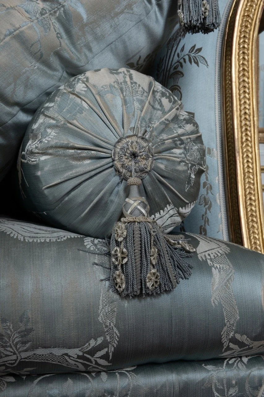 A decorative round cushion with a pleated design, silver and gray fabric, intricate embroidery, and a tassel with ornate silver charms, resting on a matching upholstered sofa with floral patterns and a gold-framed mirror in the background.