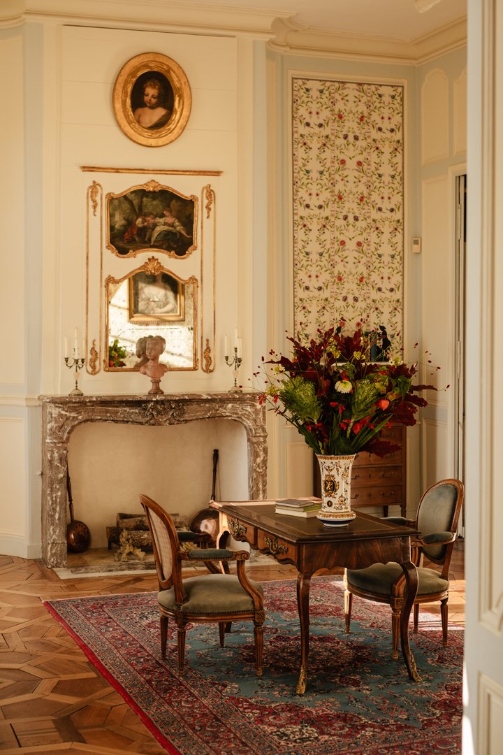 A vintage-style living room with an ornate fireplace, a large floral arrangement on a wooden table, two upholstered chairs, a Persian-style rug, and decorative wall art and mirror.