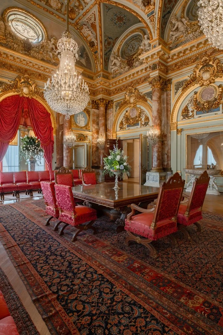 Opulent, ornate royal or aristocratic interior featuring a large wooden dining table surrounded by red velvet upholstered chairs, elaborate chandeliers, gold accents, marble columns, and rich drapery in a palace or mansion.